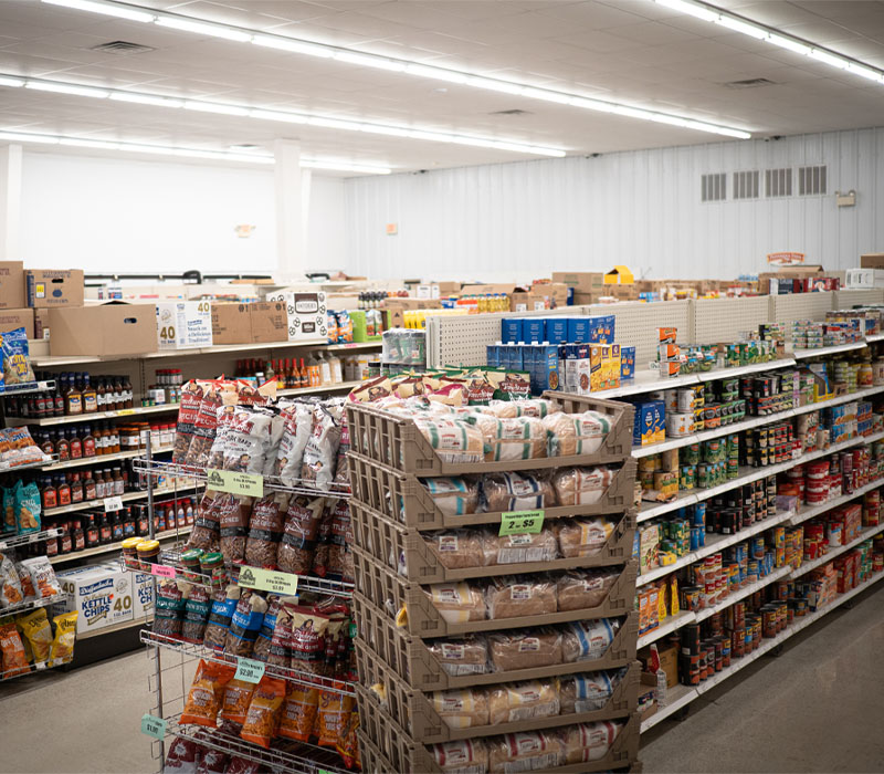 Stacked crates of bread and wire racks of snacks, aisles filled with canned and boxed items.